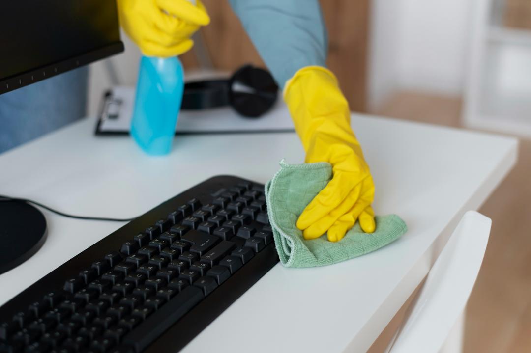 Two hands wearing yellow gloves spraying and wiping cleaning products from a desk with a computer screen and keyboard